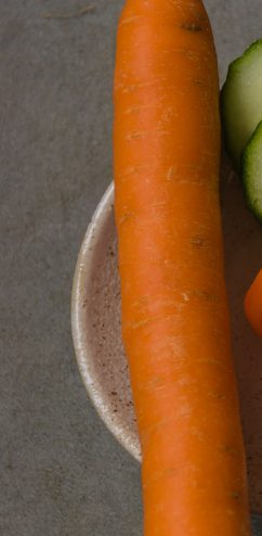Eine große, orange Karotte auf einem hellen Teller, neben einer Gurke und einer Orange.